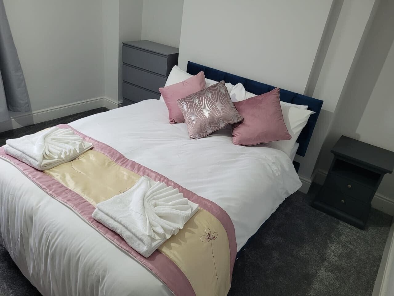 Een keurig opgemaakt bed met een wit en roze dekbedovertrek, een blauw hoofdbord en een klein nachtkastje.