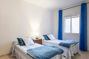Una habitación con dos camas, una mesita de noche, una ventana con cortinas y una lámpara.