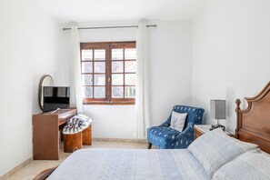 Un dormitorio con cama, una silla, un escritorio, un televisor y una ventana con cortinas.