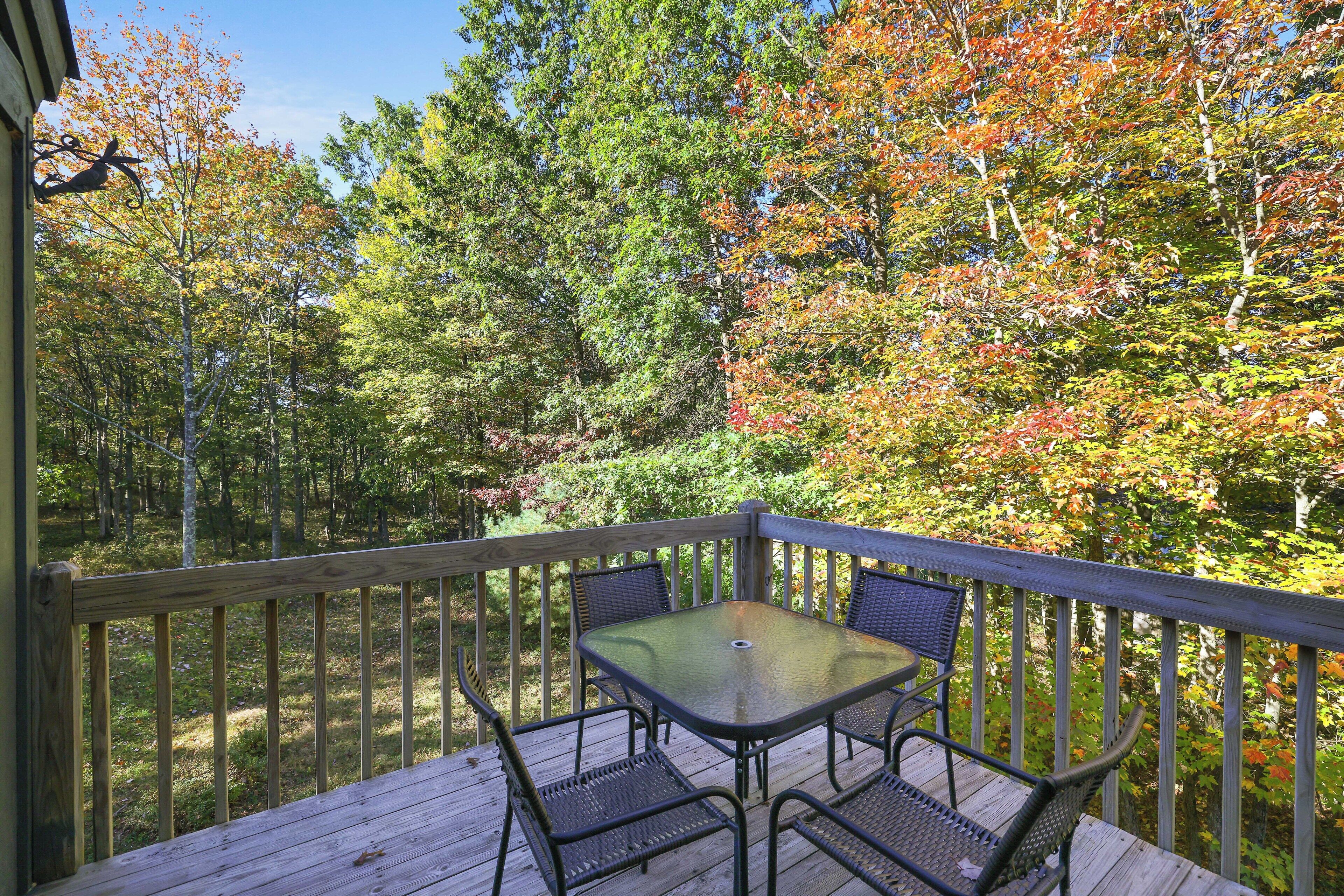 A wooden deck with a glass table and four chairs, surrounded by trees with autumn foliage.