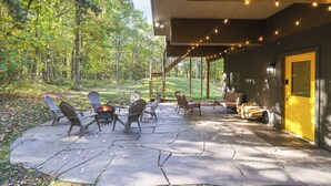 Un patio exterior con sillas Adirondack, un hogar para fogata y un área cubierta con luces colgantes.