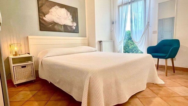 A bedroom with a bed, a nightstand, a chair, and a window with curtains. - Malea by ColombiElbaVacanze (Capoliveri)