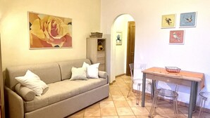 A living room with a sofa, a dining table, and chairs. There is a painting on the wall and a door leading to another room. - Malea by ColombiElbaVacanze (Capoliveri)