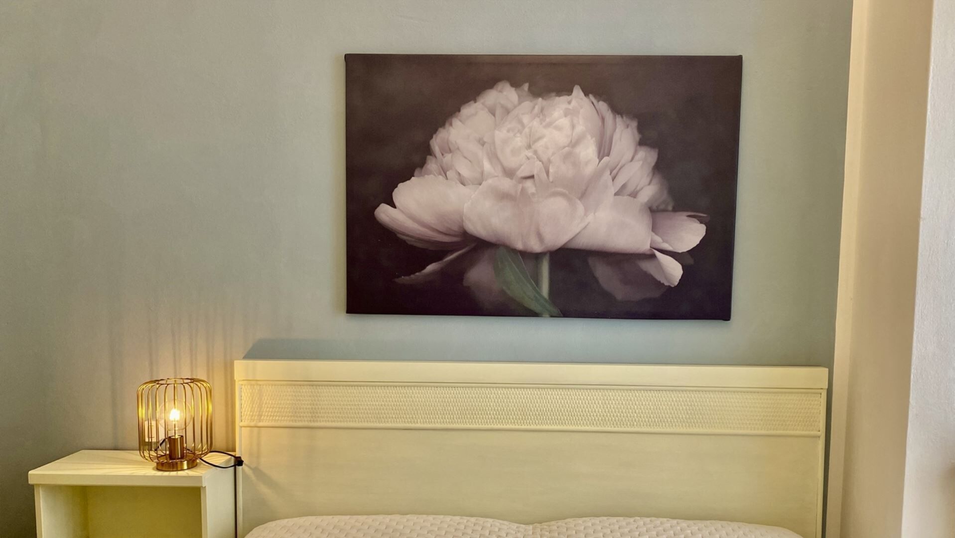 Een slaapkamer met een wit hoofdeinde, een nachtkastje met een lamp, en een bloemschilderij aan de muur.