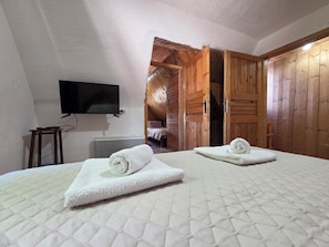 Une chambre avec deux lits, un téléviseur fixé au mur, des portes en bois et un lit visible avec une serviette roulée.