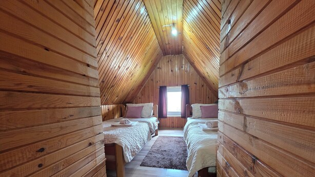 Un dormitorio abrigado en el ático con dos camas, paredes de madera, techo inclinado y una ventana con cortinas.