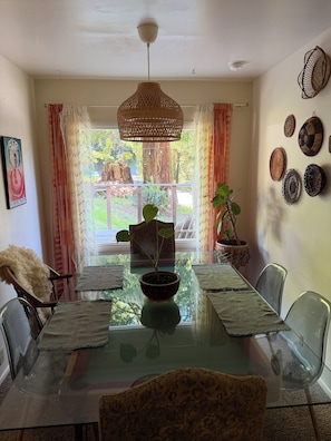 Un coin repas avec une table en verre, des chaises, un luminaire suspendu et une vue sur les arbres à travers les rideaux.