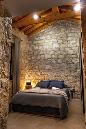 A bedroom with stone walls, a wooden ceiling, a bed with dark bedding, and a small bedside table with a lamp.