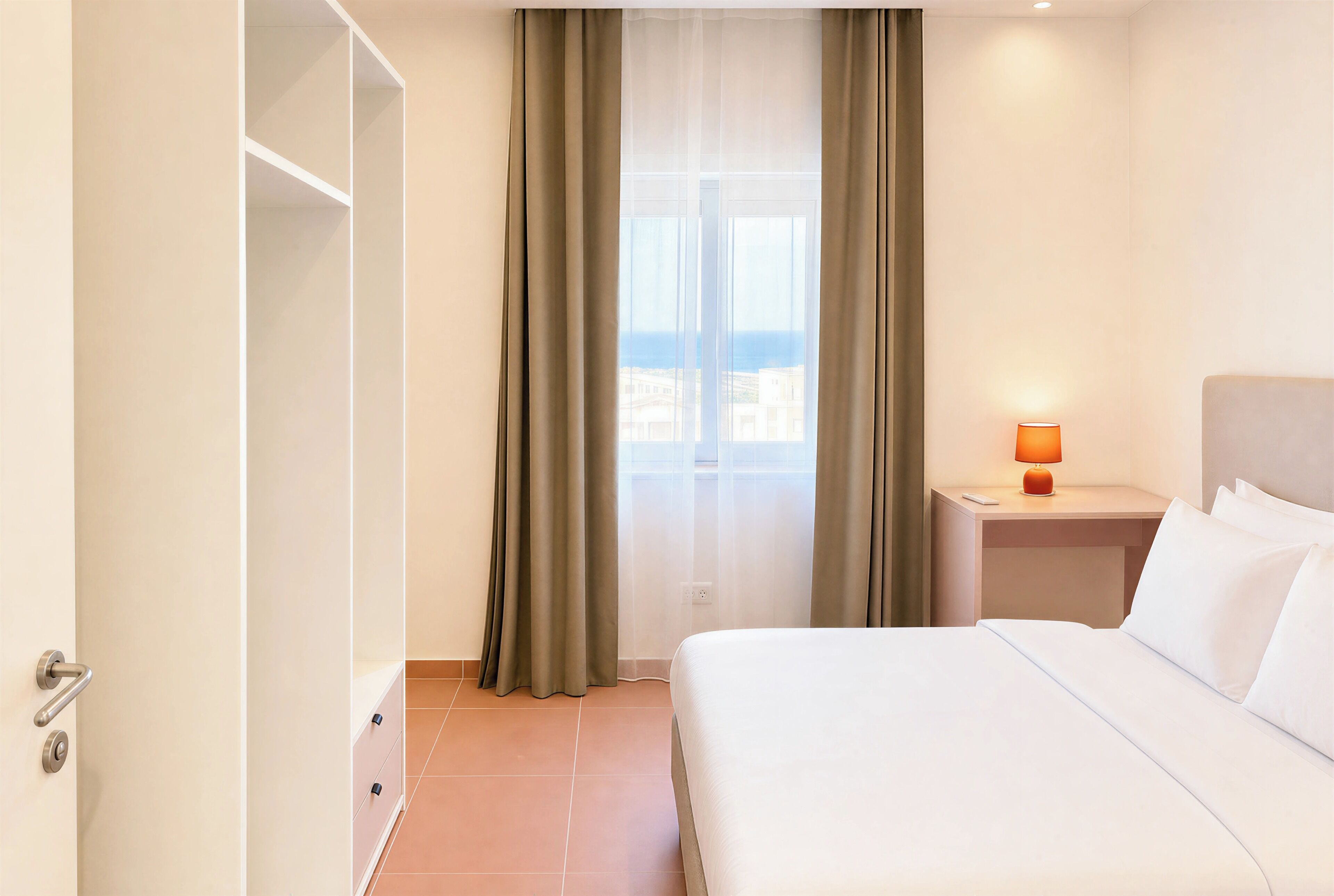 Habitación de hotel con cama, mesita de noche con lámpara, ventana con cortinas y vista a la ciudad.