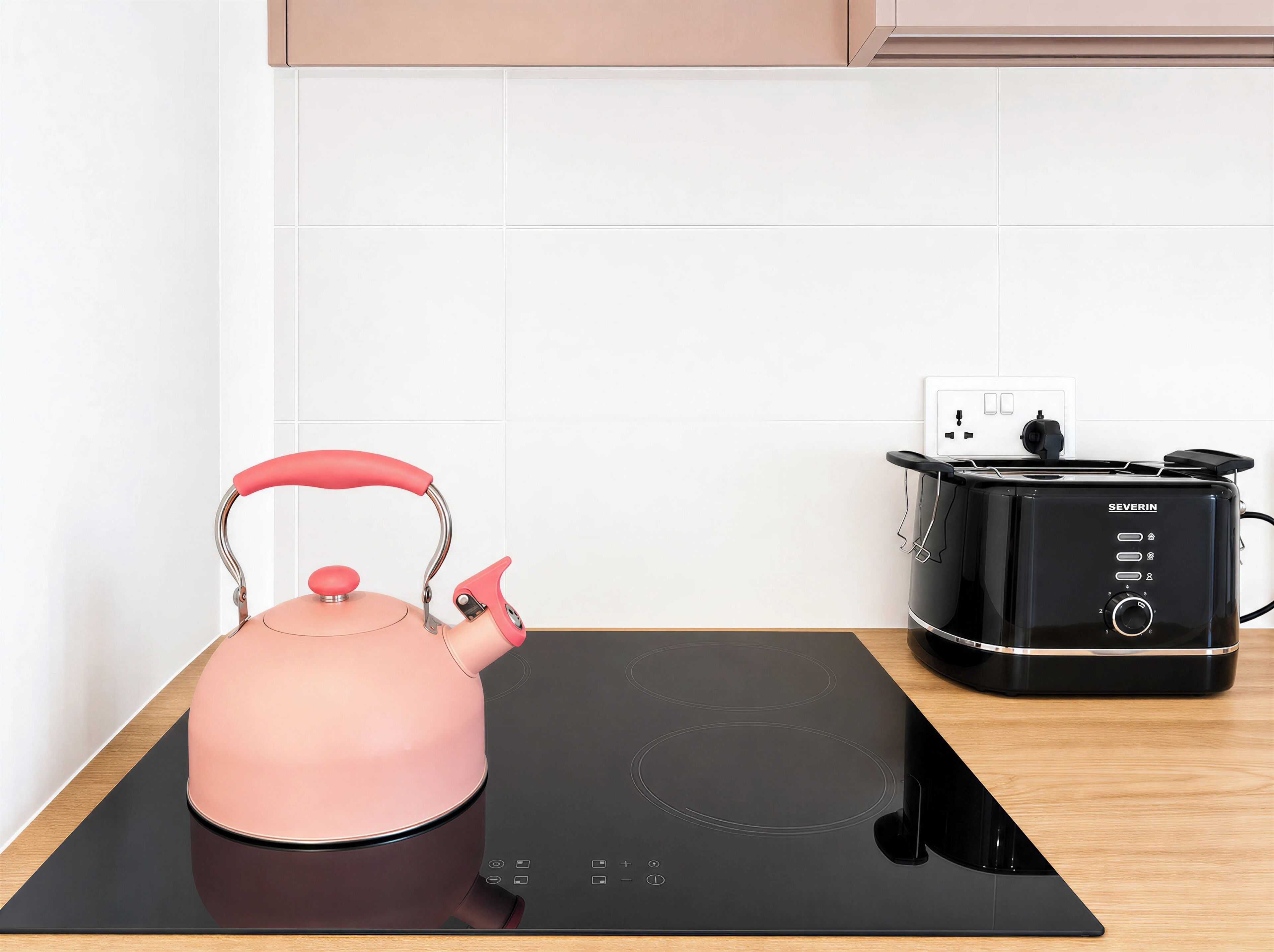 A pink kettle on a black induction cooktop, a black toaster oven, and a white wall with a power outlet.