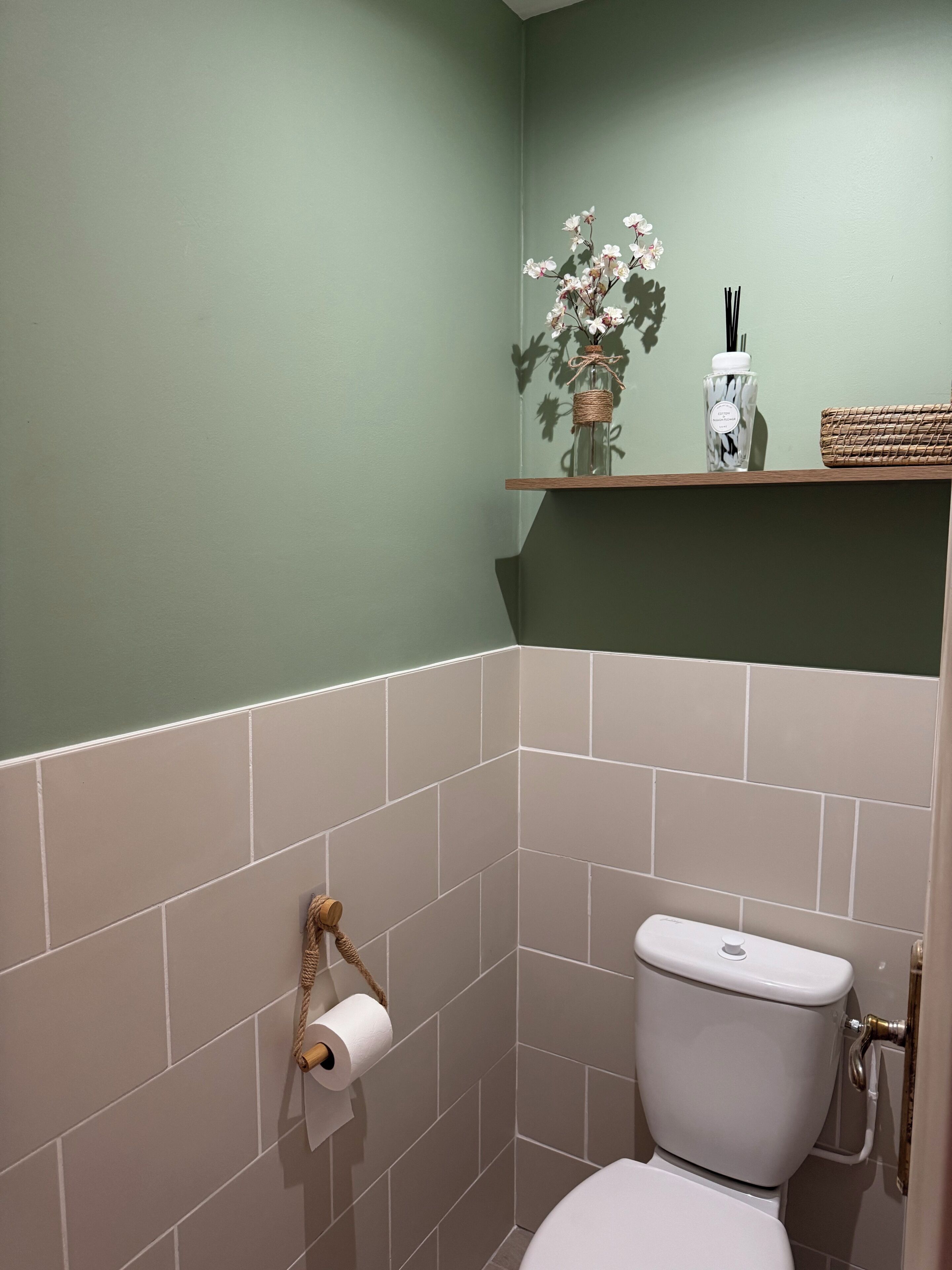 Une salle de bain avec des toilettes, un rouleau de papier toilette, une petite étagère avec un vase de fleurs et un flacon de gel hydroalcoolique.