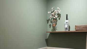 A bathroom with a toilet, a roll of toilet paper, a small shelf with a vase of flowers, and a bottle of hand sanitizer.