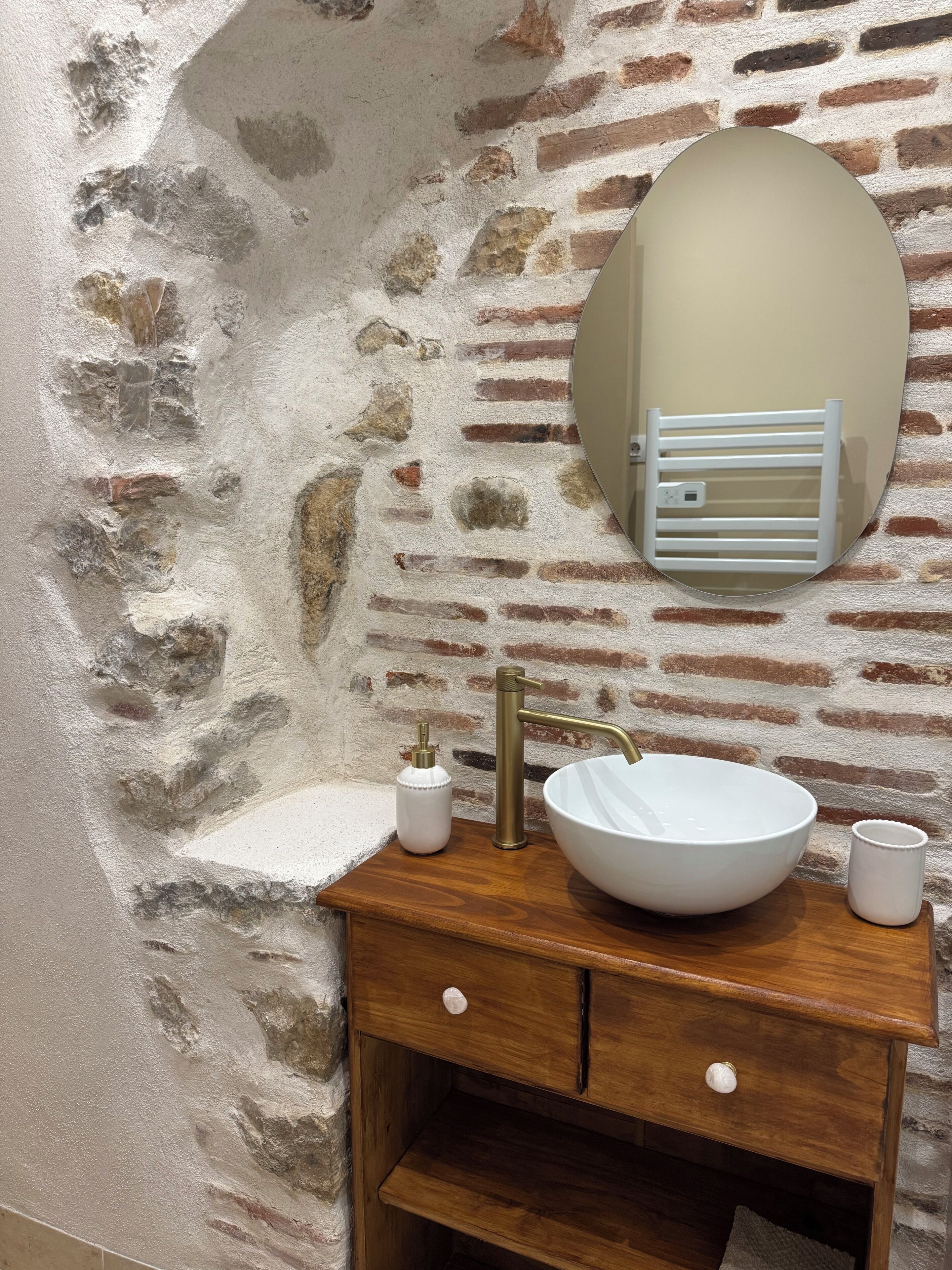 A bathroom with a stone wall, a wooden vanity, a white sink, a brass faucet, a mirror, and a towel rack.