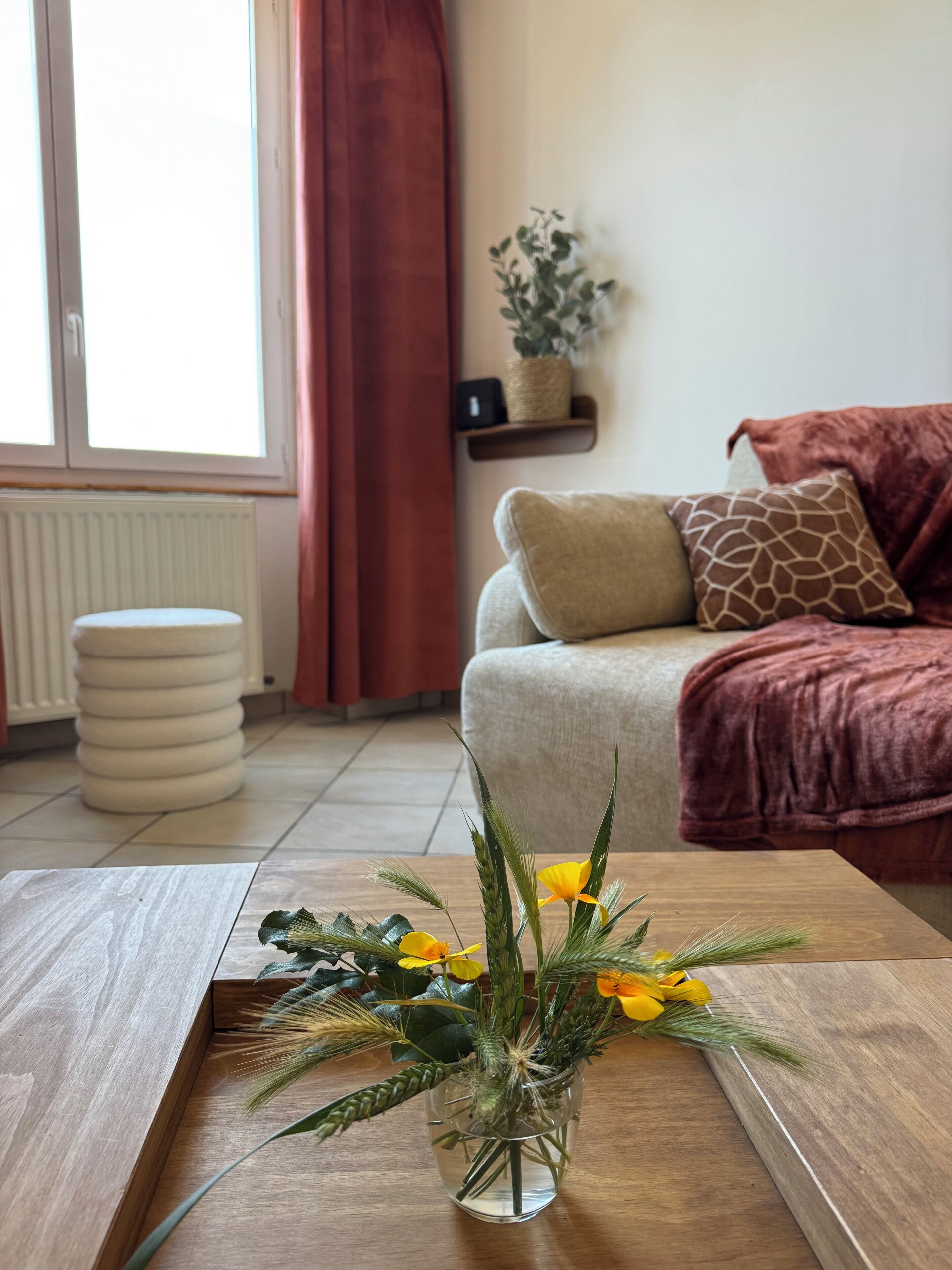Una sala de estar con una mesa de centro de madera, un sofá con cojines, una planta en maceta y un ventanal con cortinas.