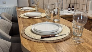 A wooden dining table set with plates, cutlery, and glasses, with a kitchen in the background. - Cosy apartment on the banks of the Nivernais Canal (Saint-Léger-des-Vignes)