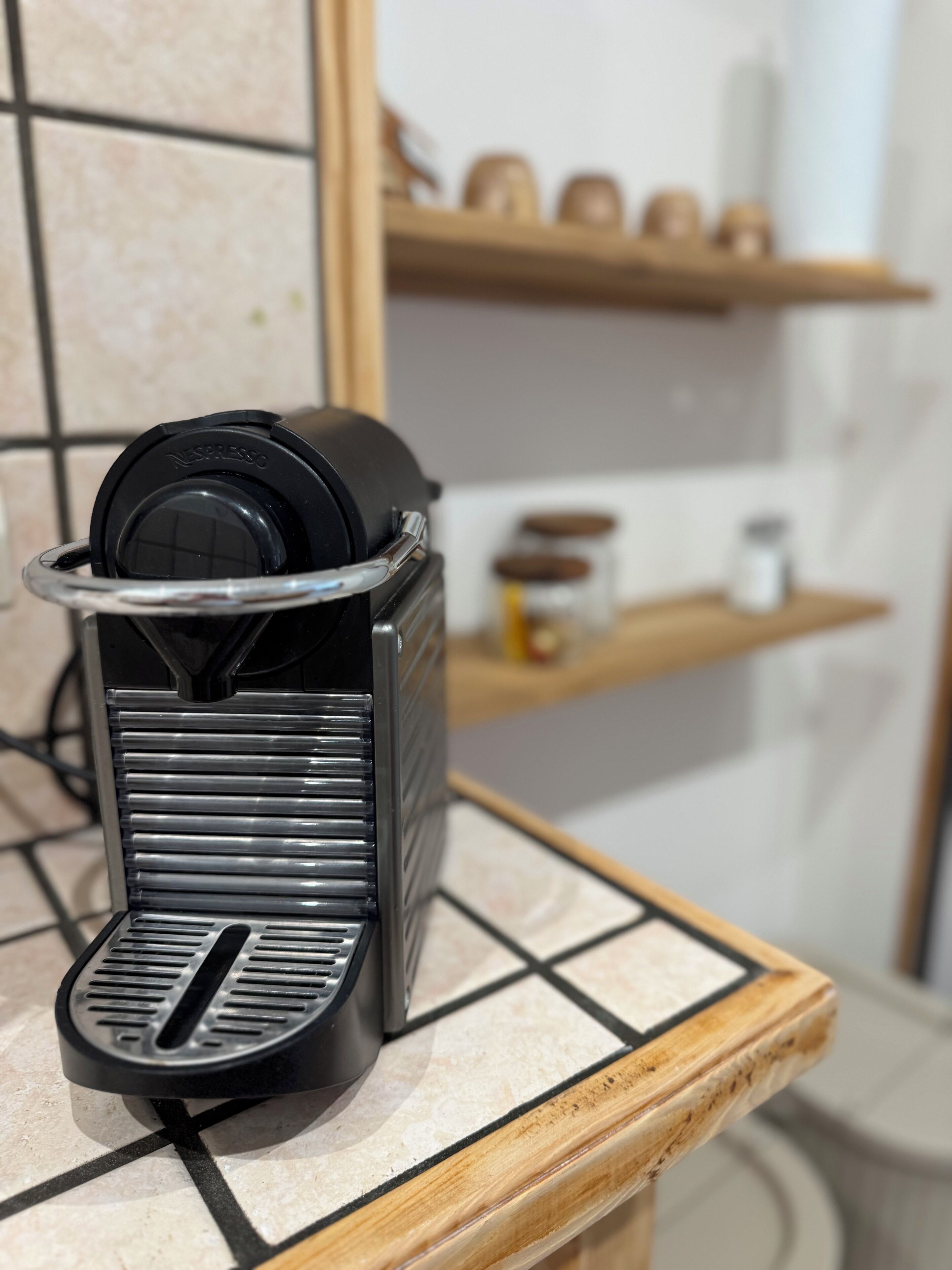 Kahvinkeitin keittiön työtasolla, taustalla laatoitettu seinä ja puiset hyllyt.