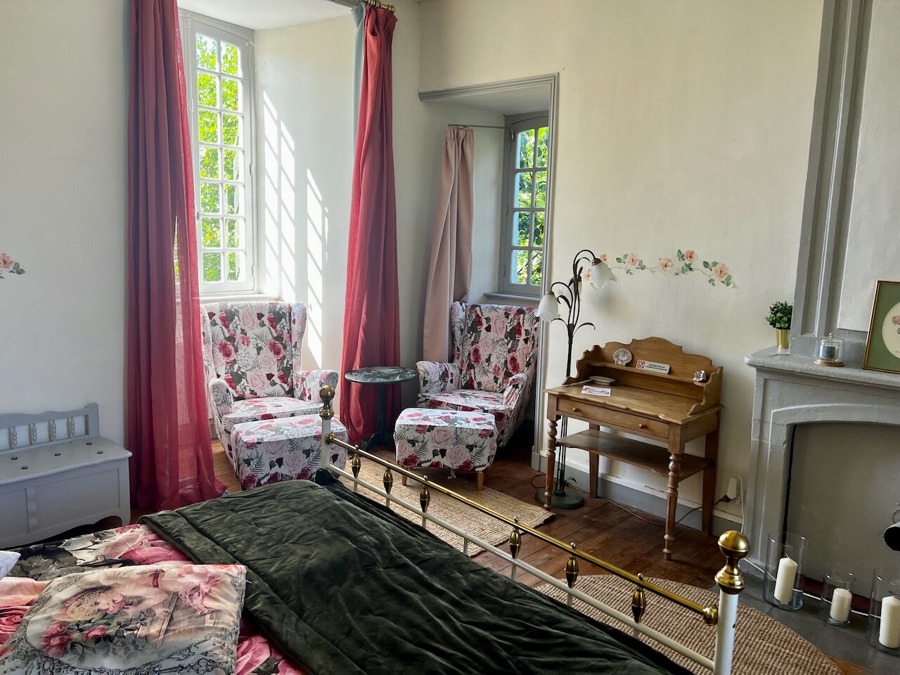 A bedroom with a bed, two chairs, a desk, a mirror, and a fireplace.