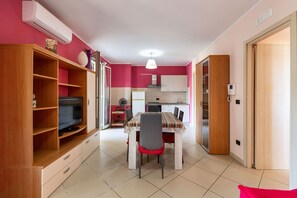 Ein gemütlicher Wohnraum mit Esstisch, Stühlen, einem Fernseher auf einem Holzregal, einer Küchenzeile und einer Tür zu einem weiteren Zimmer.
