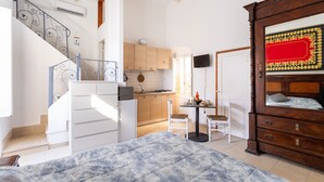 Un dormitorio con cama, cómoda de madera, mesa de comedor con sillas, zona de cocina y escalera.