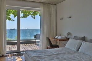 A hotel room with a bed, a desk with a chair, a table with towels, and a view of the sea through a sliding door.