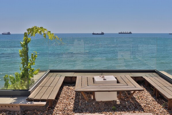 Ein Holzdeck mit Tisch und Bänken mit Blick auf ein ruhiges Meer, in der Ferne Schiffe.