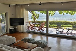 Ein Wohnzimmer mit Fernseher, Sitzgelegenheiten und einer großen Schiebetür zu einer Terrasse mit Gartenmöbeln.