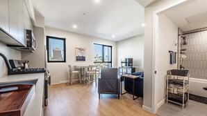 Een moderne keuken met een eethoek, een grijze fauteuil en uitzicht op de stad door een groot raam.