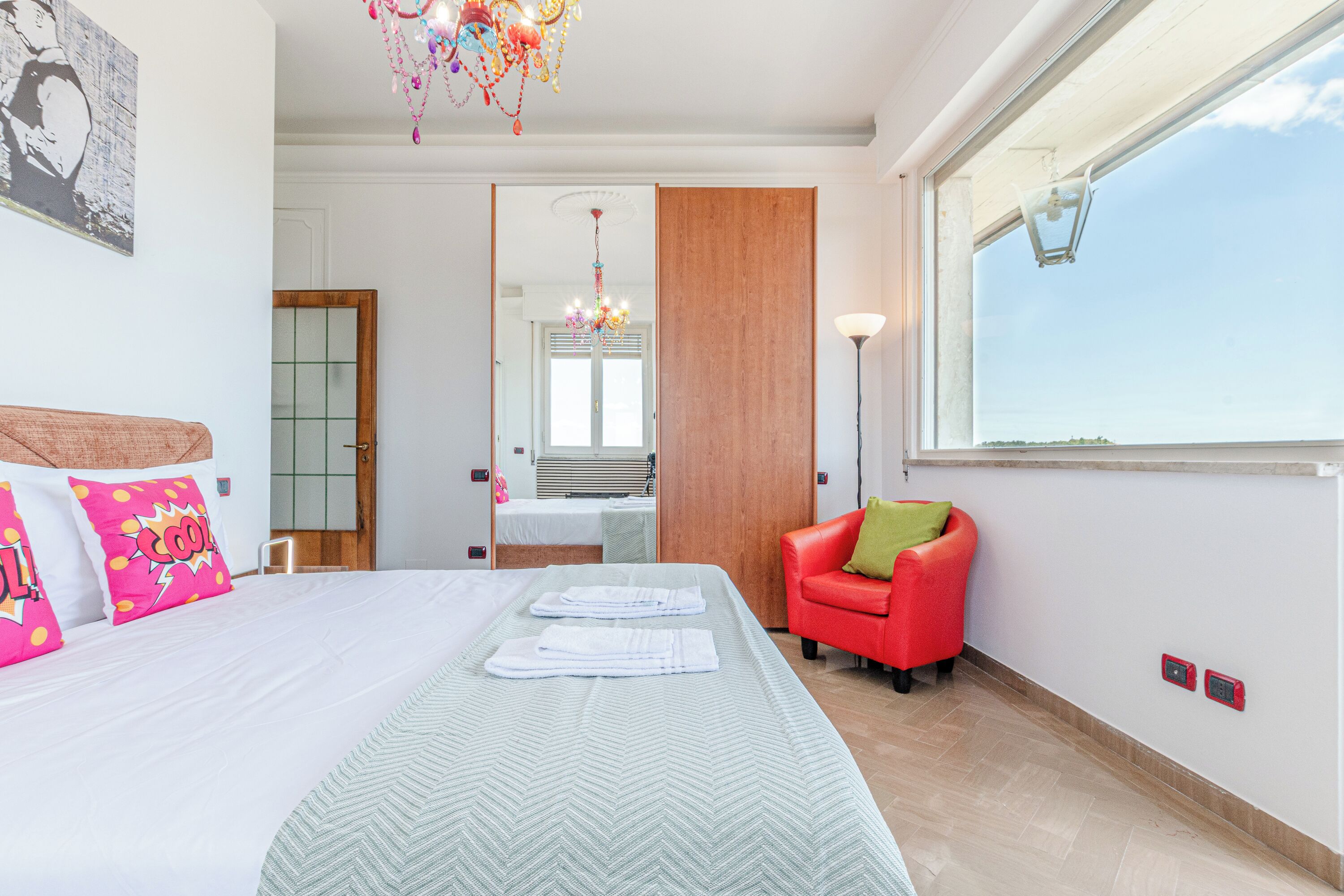 A hotel room with a bed, a red chair, a window, and a chandelier.