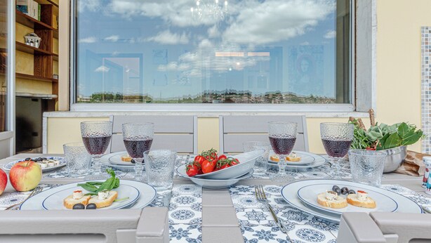 Ein Esstisch ist gedeckt mit Tellern, Gläsern und einer Tomatenschale, mit Blick auf den Himmel durch ein Fenster.