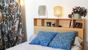 Een bed met een bedsprei met dessin, een nachtkastje met spiegel, een lamp en een plank met diverse spullen.