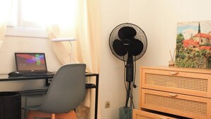 Een kamer met een bureau, stoel, computer, ventilator en een schilderij aan de muur.