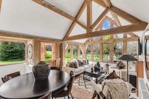 A spacious room with a high ceiling, wooden beams, a dining table, sofas, and large windows offering a view of the outdoors. - The Subedge Collection - A Unique Cotswolds Escape (Weston Subedge)