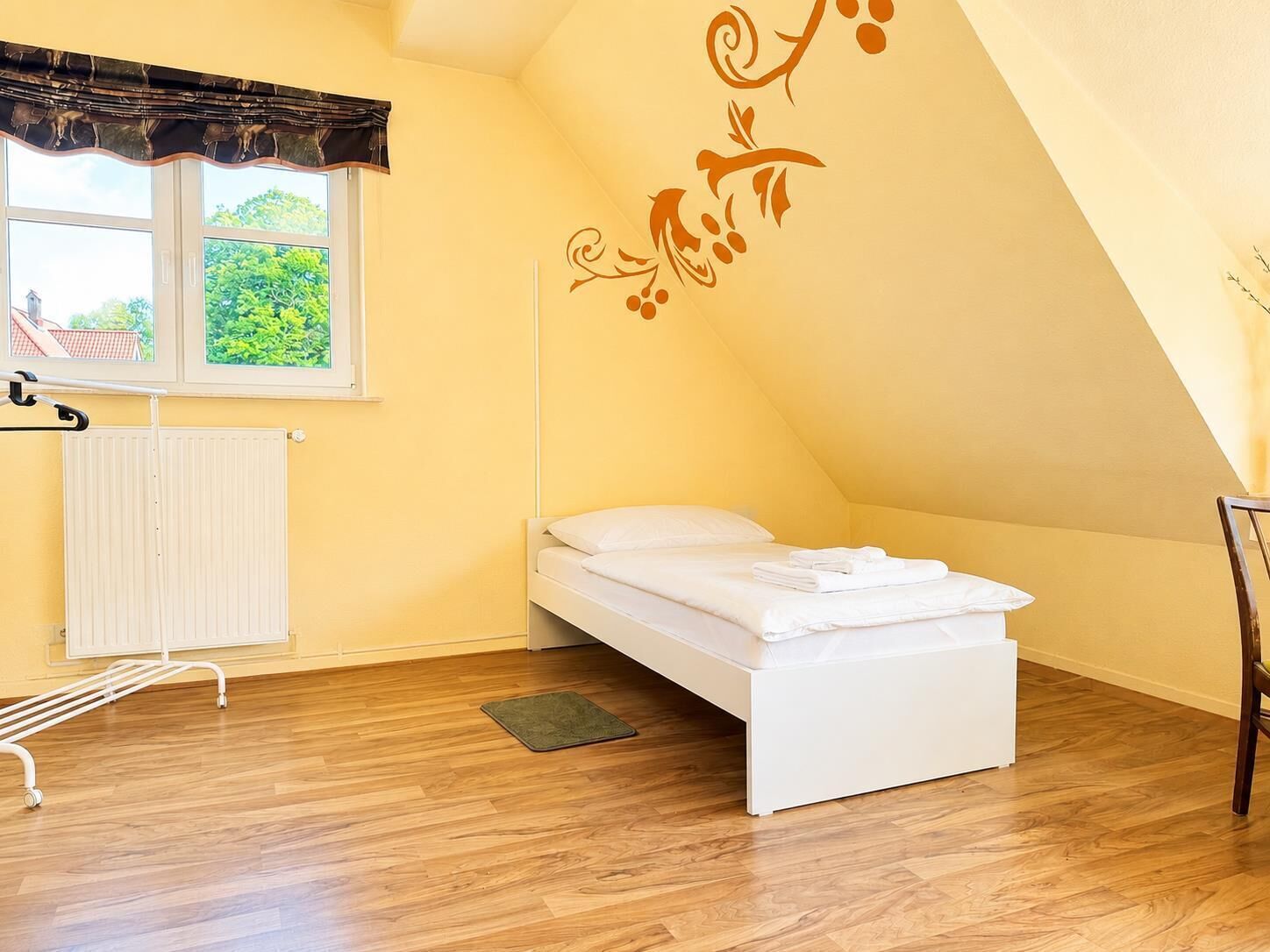 Une petite chambre avec un lit simple, une fenêtre, un radiateur et un motif décoratif sur le mur.
