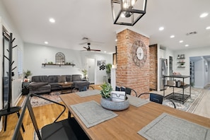 A modern living room with a dining area, a brick accent wall, and a ceiling fan. - The Glow-Up House | Pool, Spa & Glam Room (Phoenix)