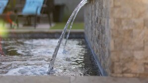 Una fuente de agua con un muro de piedra, un patio con sillas y una sombrilla.