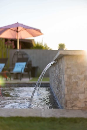 A water feature with a stone wall, a patio area with chairs, and an umbrella. - The Glow-Up House | Pool, Spa & Glam Room (Phoenix)