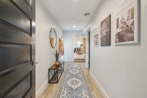 A hallway with a dark wooden door, a mirror, a bench with a plant, a patterned rug, and framed pictures on the wall. - The Glow-Up House | Pool, Spa & Glam Room (Phoenix)