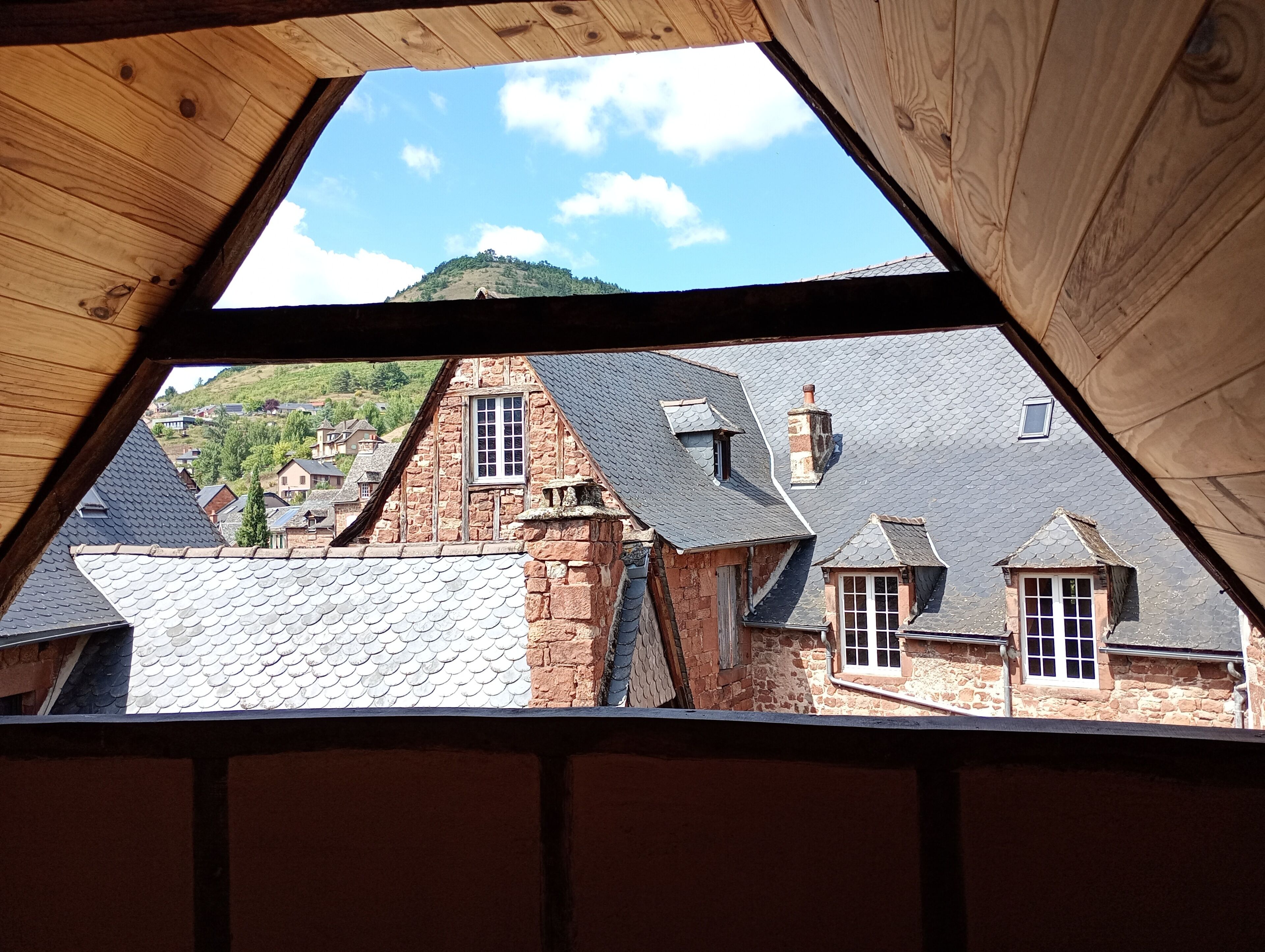 A view through a triangular window of a building with stone walls and slate roofs, overlooking a hillside with other buildings.