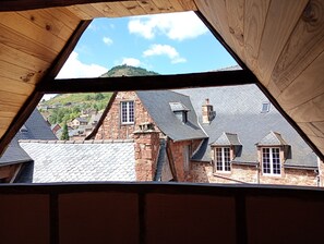 Une vue à travers une fenêtre triangulaire d’un bâtiment aux murs de pierre et aux toits d’ardoise, donnant sur une colline où se trouvent d’autres bâtiments.