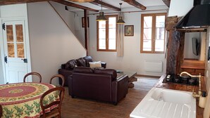 A rustic interior with exposed wooden beams, a sofa, a dining table with a patterned tablecloth, and a kitchen area with a sink and stove. - Gite Le Mansois (Marcillac-Vallon)