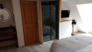 A bedroom with a wooden door, a glass wardrobe, a bedside table, a bed, a wall-mounted TV, and a standing fan. - Gite Le Mansois (Marcillac-Vallon)