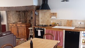A rustic kitchen with a wooden table, chairs, a gas stove, and a black range hood. - Gite Le Mansois (Marcillac-Vallon)