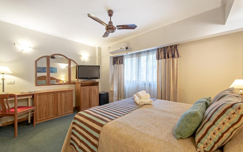 A hotel room with a bed, a desk with a television, a ceiling fan, and a window with curtains.