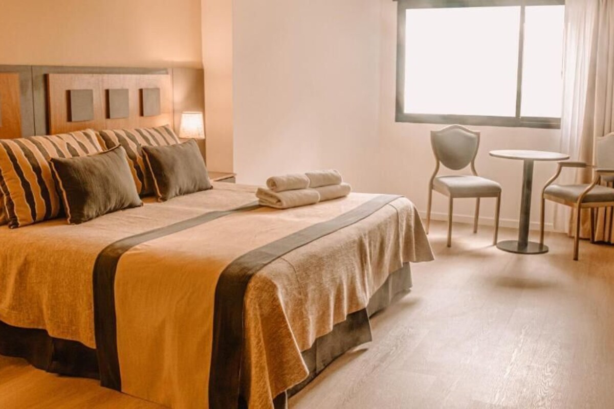 Quarto de hotel com uma cama grande, duas cadeiras, uma mesinha e uma janela.
