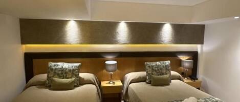 Habitación de hotel con dos camas individuales, un cabecero de madera, lámparas de noche y una mesita de noche pequeña.