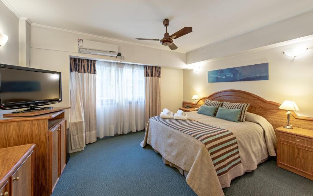 Une chambre d’hôtel avec un lit, une télévision, un ventilateur de plafond, une table de chevet, une lampe et un tableau au mur.