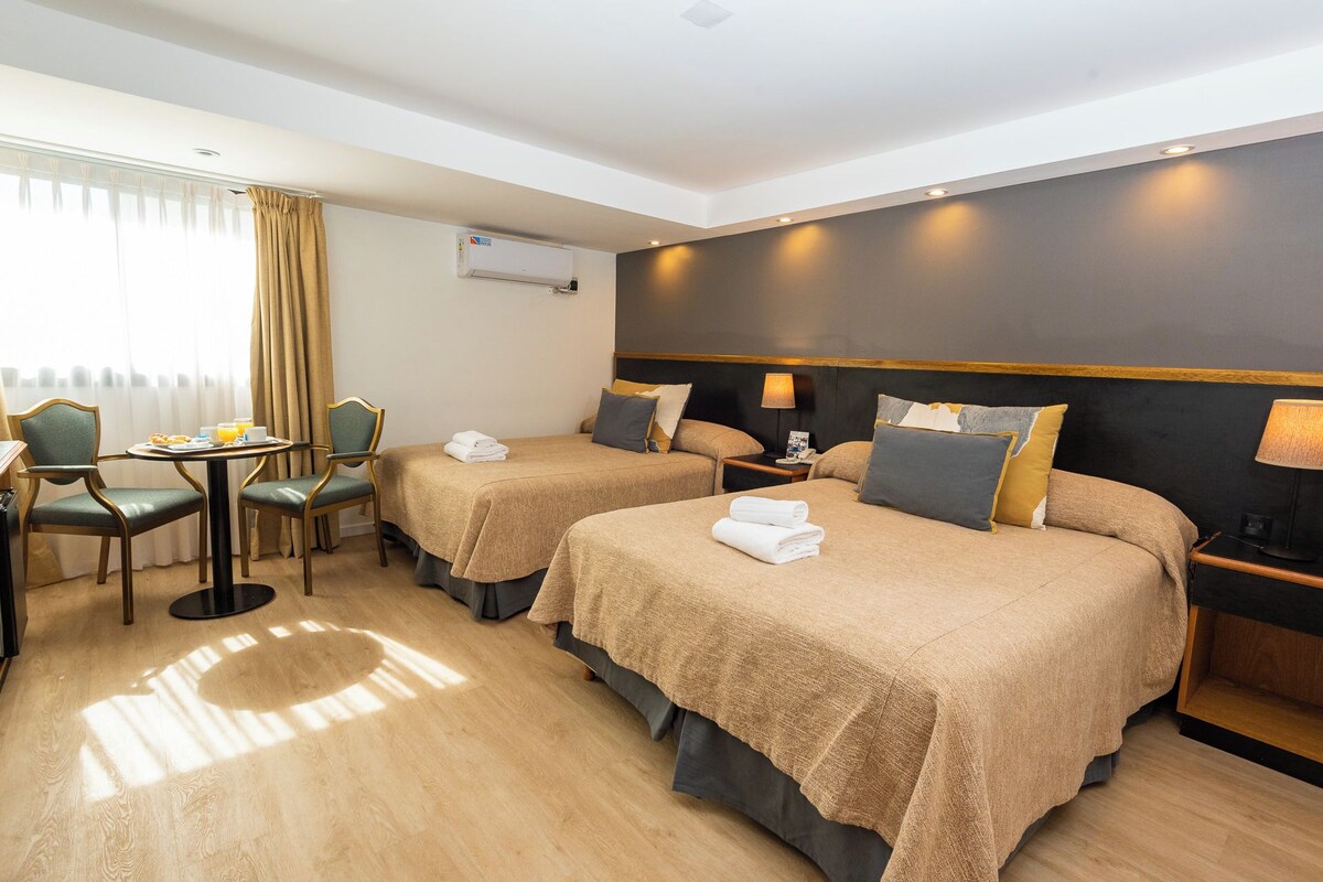Quarto de hotel com duas camas, área de refeições e janela com cortinas.