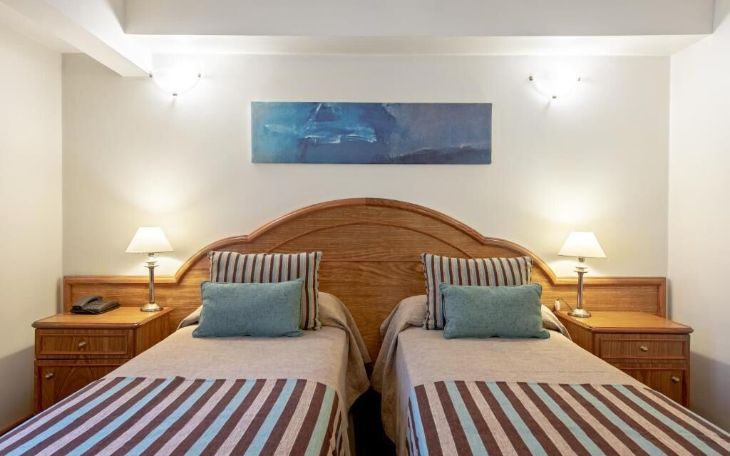 Quarto duplo com duas camas, luminárias de cabeceira, um painel de madeira na cabeceira e um telefone na mesinha de cabeceira.
