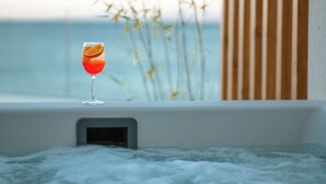 A glass of red drink with an orange slice on a windowsill, a hot tub, and a view of the sea.