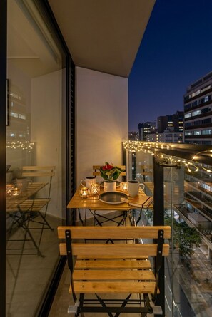 A balcony with a wooden bench, a table with candles, and a view of the cityscape at dusk. - 2BR/2BA w/ View · A/C · Pool · 24/7 Doorman (Miraflores)